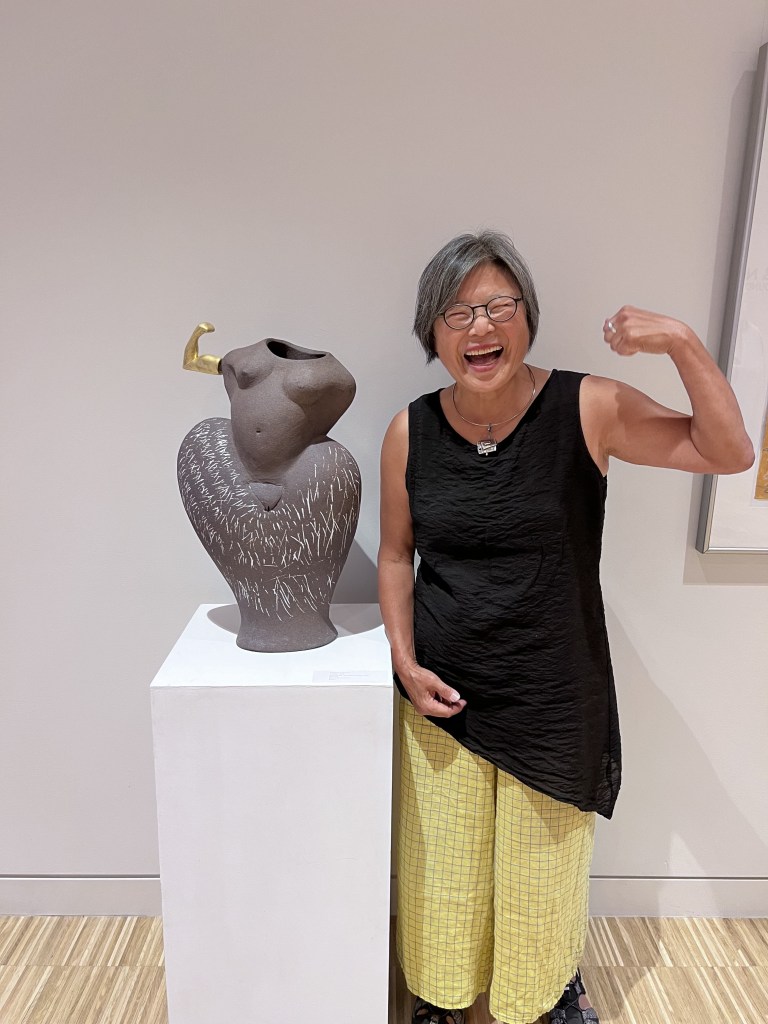 Stoneware ceramic torso sculpture with one gold arm making a fist in a triumphant "make a muscle" pose, with an older Asian woman standing next to it copying the pose. 